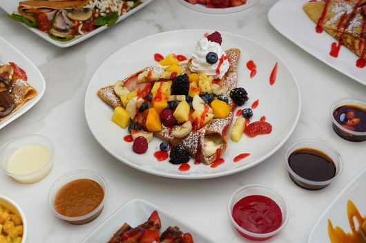 Dessert crepe topped with berries and fruit on a white table with assorted sauces and plates