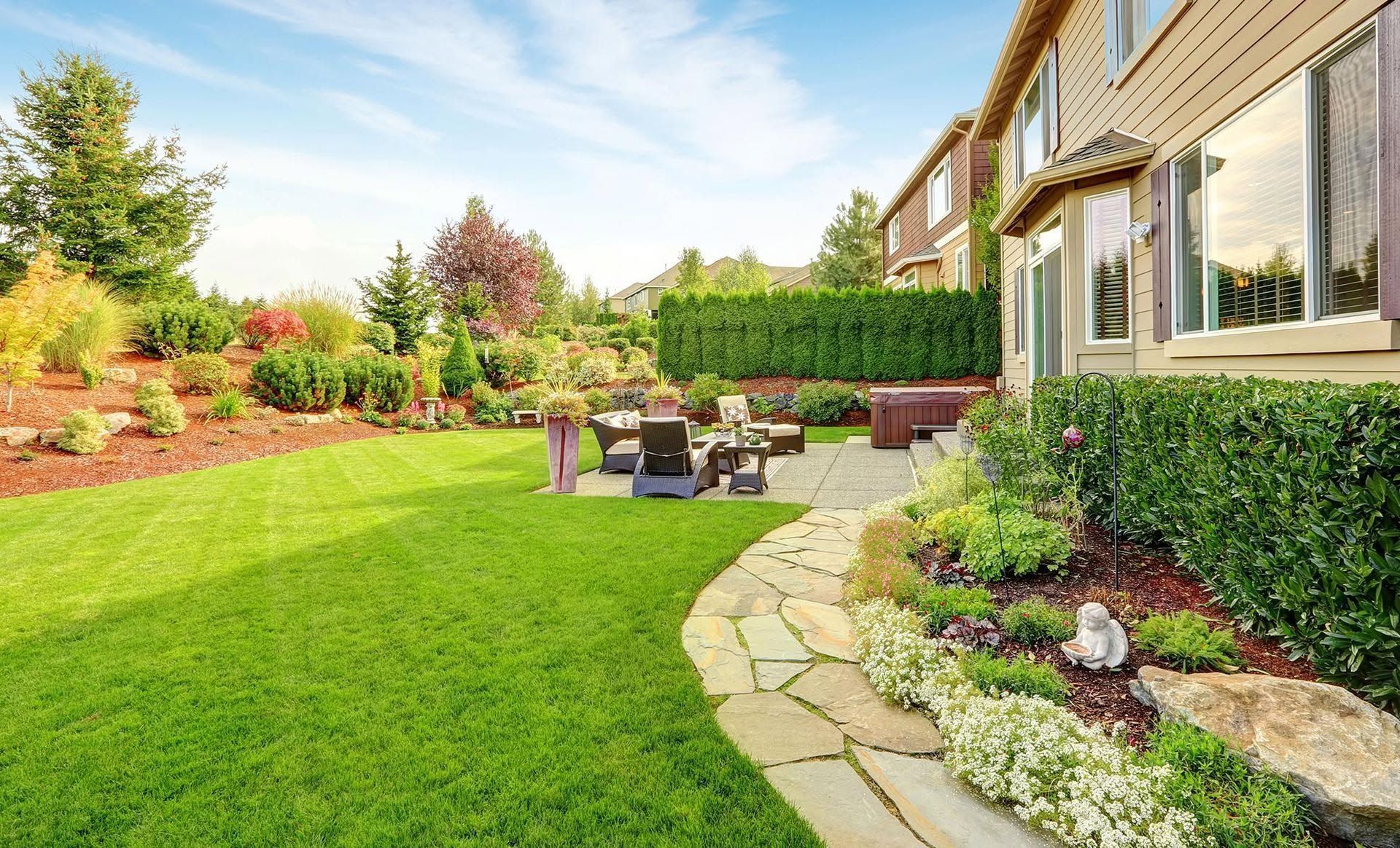 Backyard patio with lawn, hedges, stone path, and outdoor seating beside a house