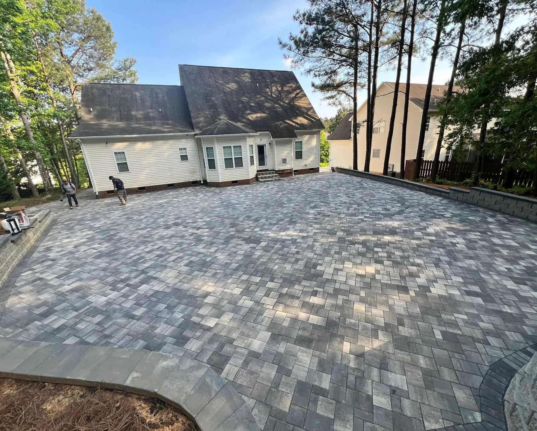 Large paver driveway leading to a light house with a dark roof and trees along the edge