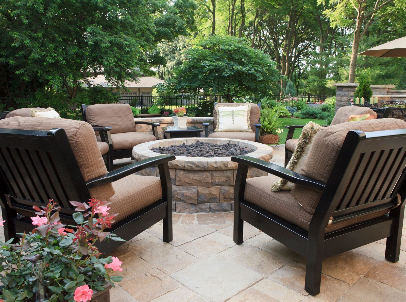 Modern backyard fire pit surrounded by white Adirondack chairs on a green lawn with a stone retaining wall.