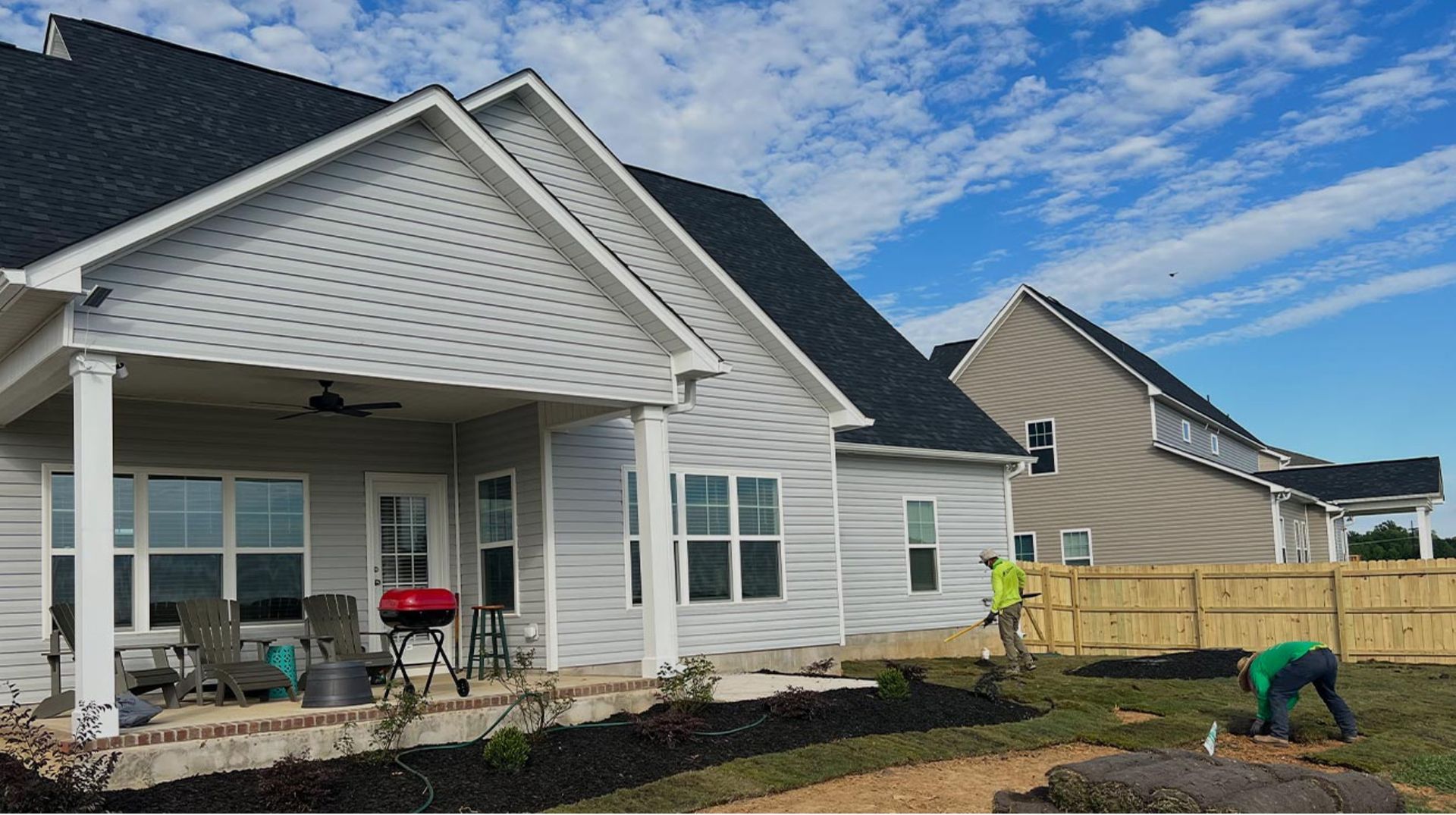 New white house with porch and black roof; workers are landscaping in the yard.