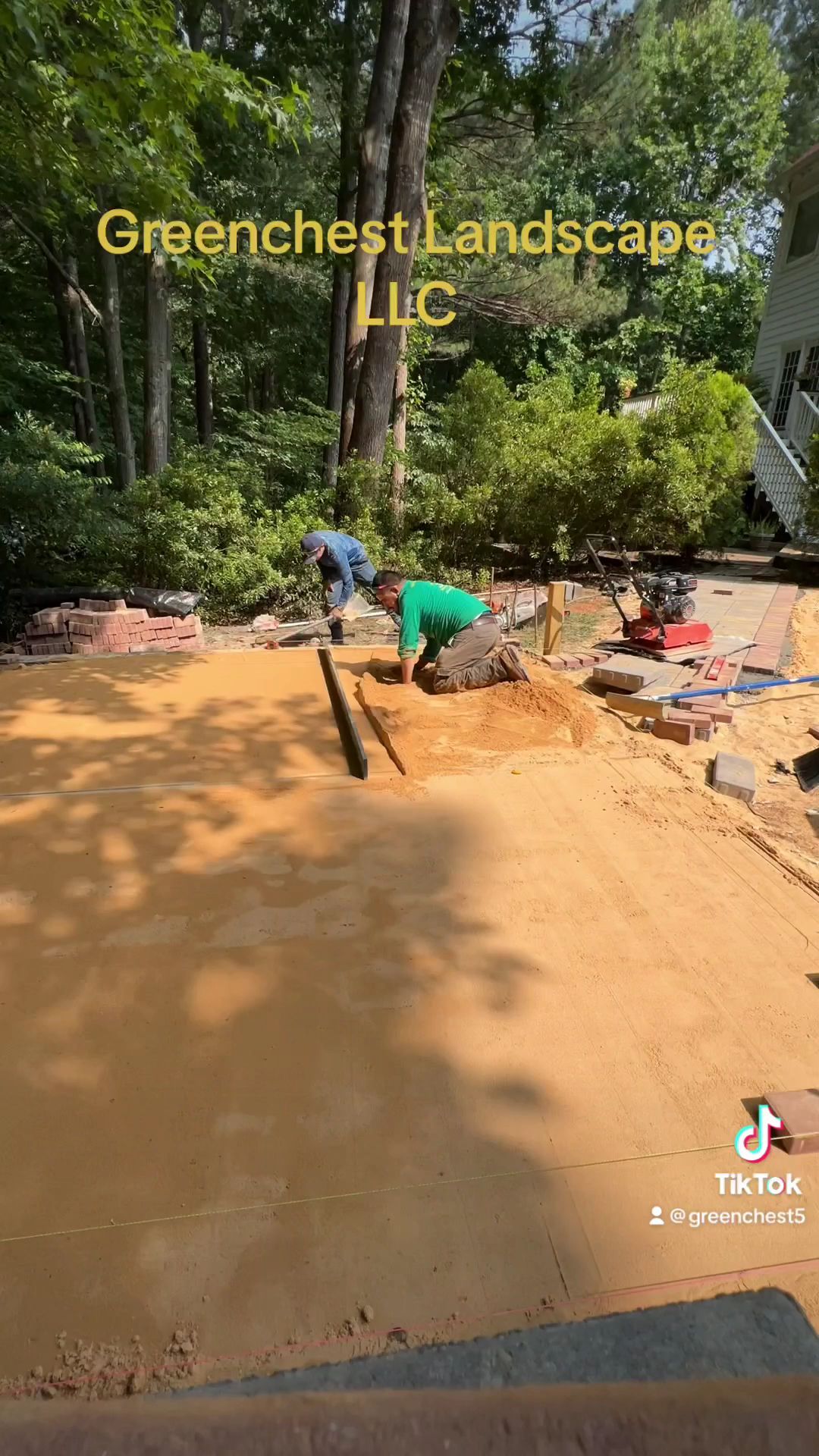 Worker smoothing tan sand in a wooded yard with landscaping materials nearby