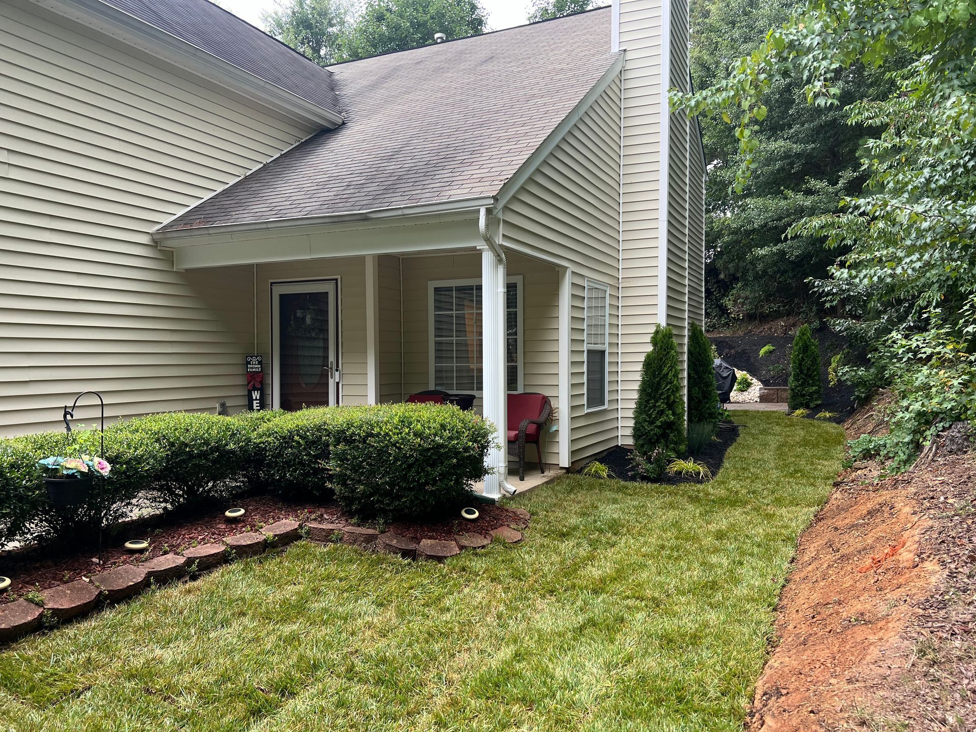 Suburban house entry with porch, shrubs, and sloped roof beside a grassy yard and wooded area