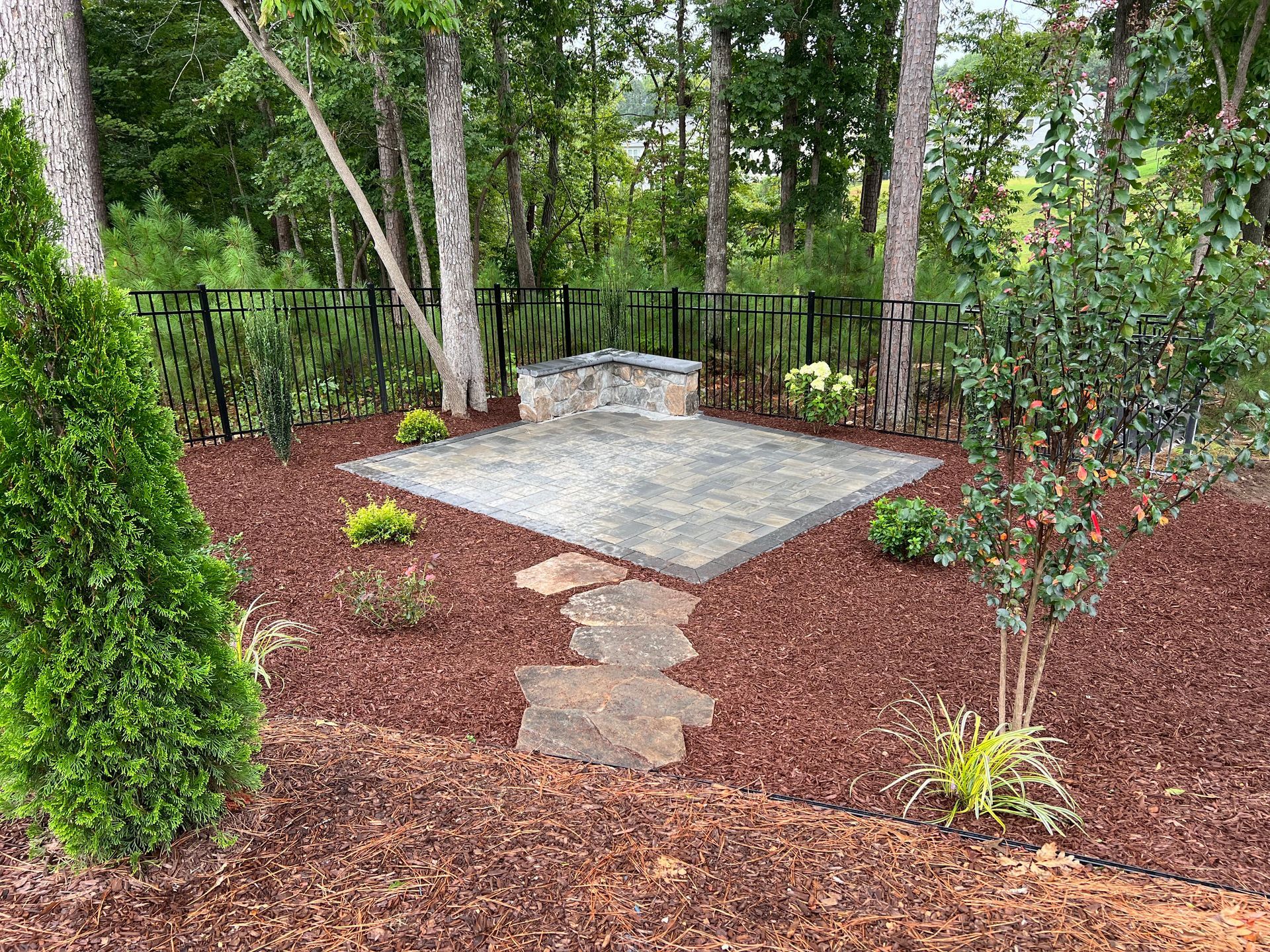 Backyard garden patio with stone slab seating area, mulch beds, trees, and black metal fence