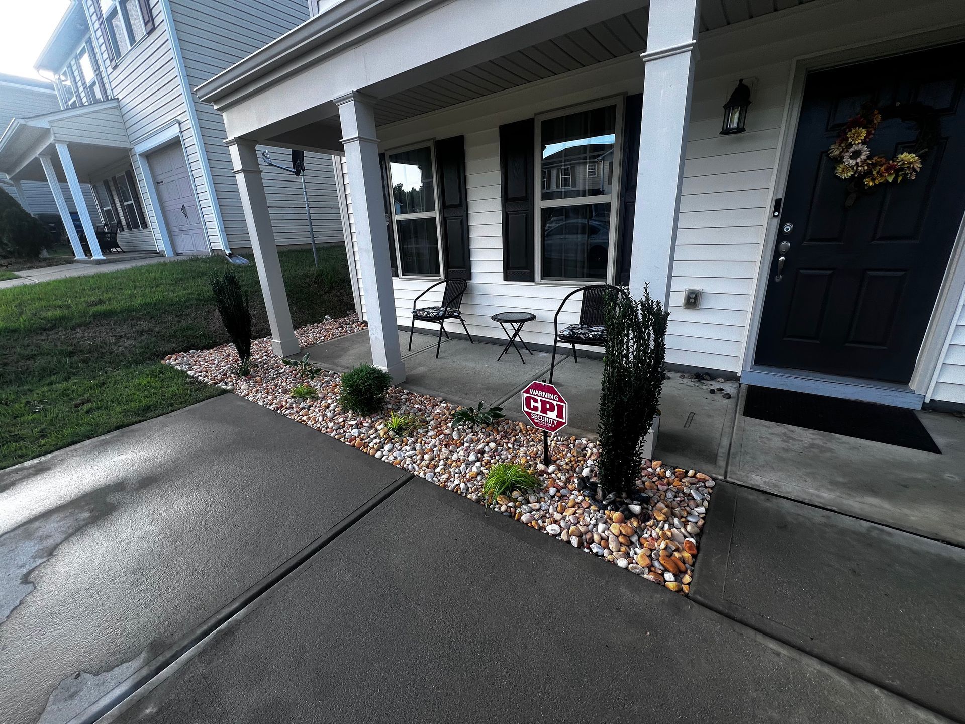 Front porch with wet concrete, white siding, black door, chairs, and a small rock garden border.