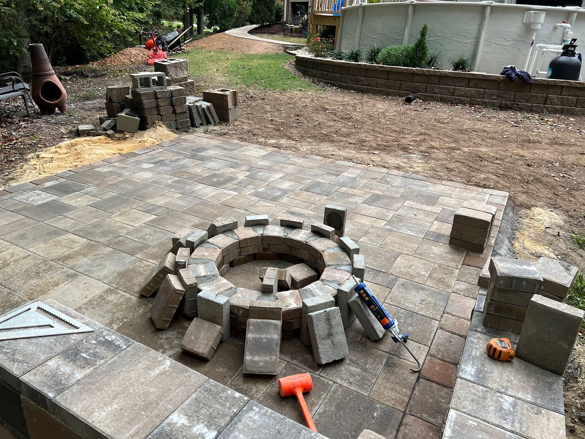 Backyard patio with circular stone fire pit under construction, surrounded by loose pavers and tools.