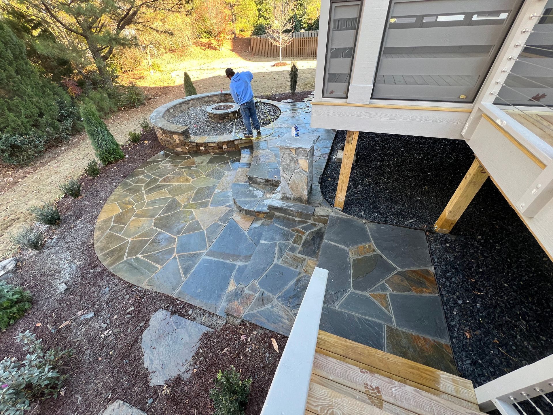 Backyard stone patio with a person near a circular fire pit and raised deck stairs