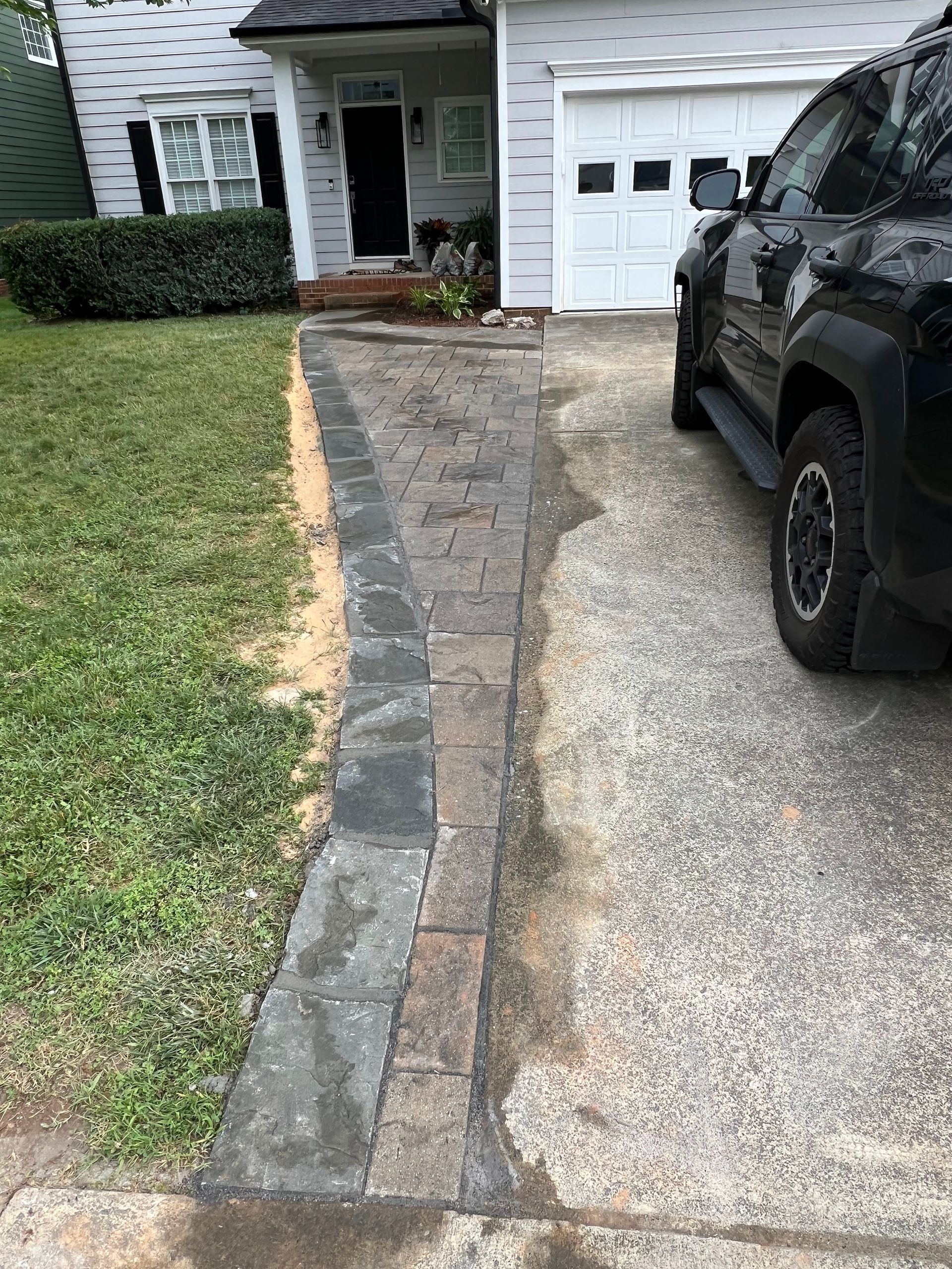 Cracked walkway beside a driveway leading to a house, with a black SUV parked on the right.