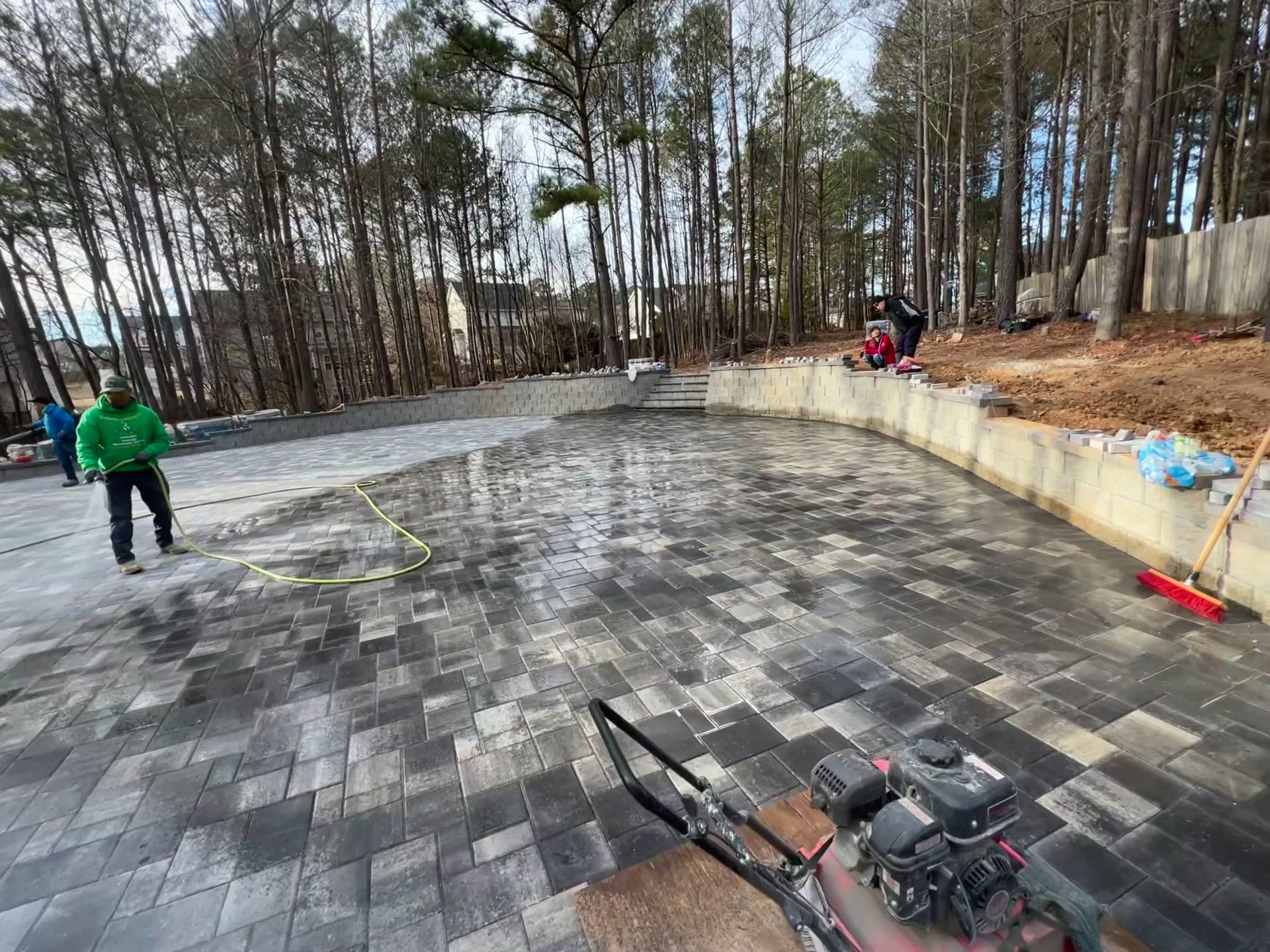 Workers installing gray pavers on a curved outdoor patio beside trees and a retaining wall
