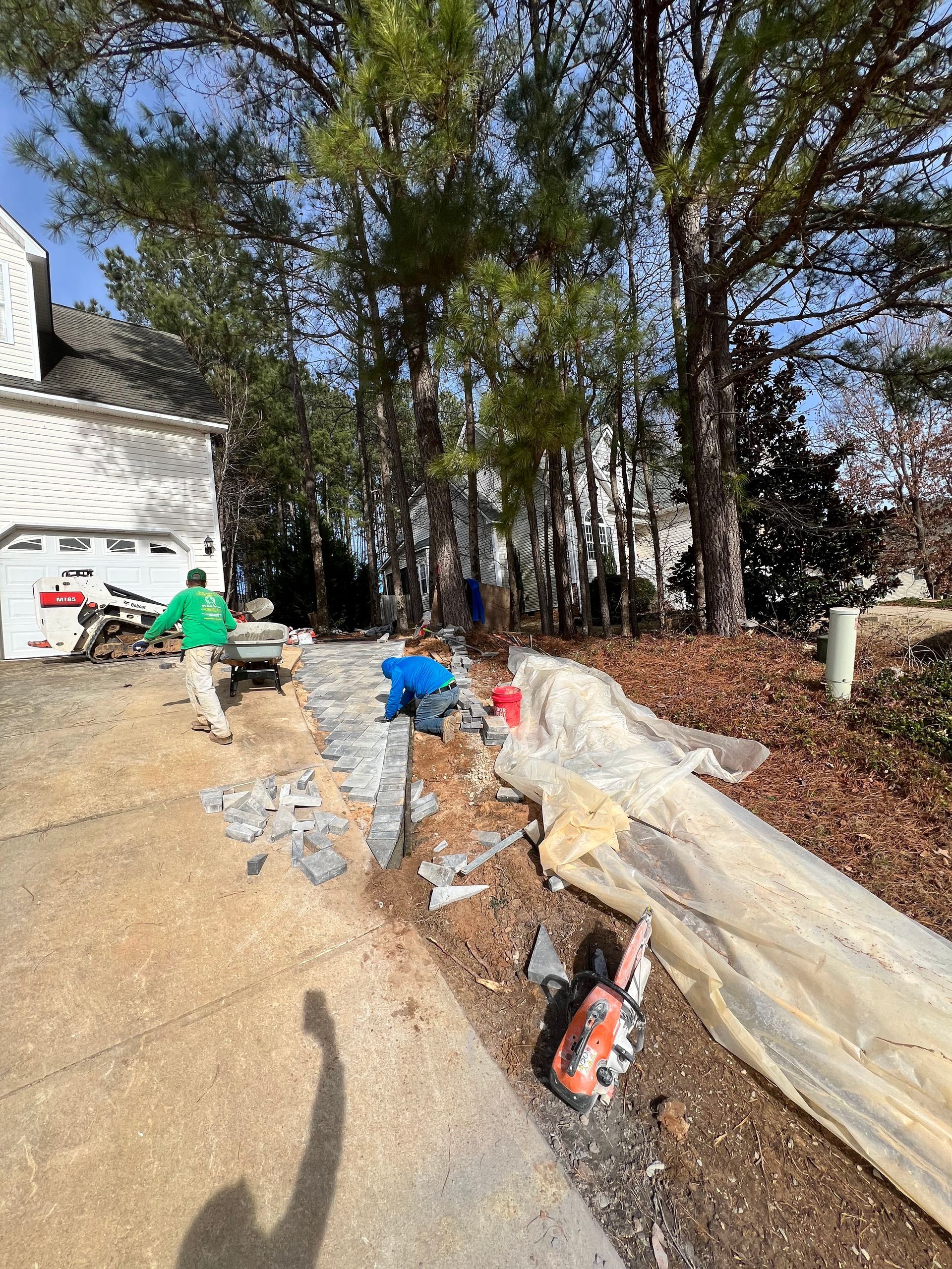 Workers laying plastic sheeting and tools along a gravel path beside a house and trees.