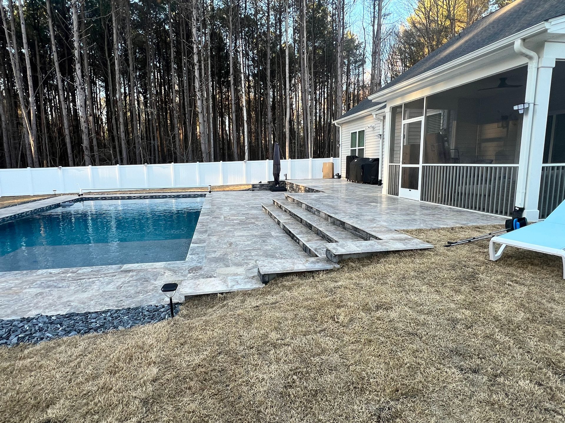 Backyard pool beside a covered patio, stone steps, and a fenced yard with tall trees in the background