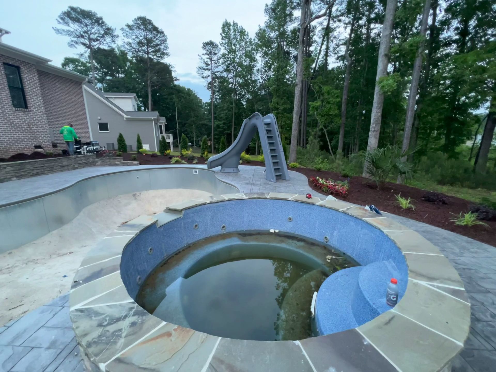 Backyard pool with a round sunken hot tub and sliding board, surrounded by trees and a house