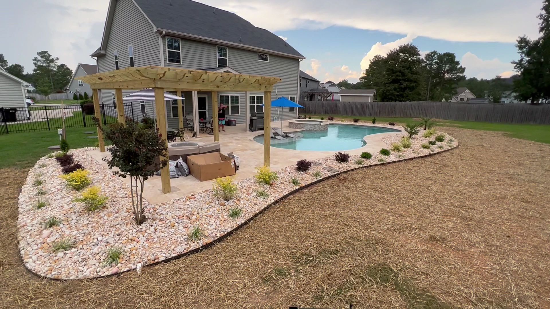 Backyard with a covered pergola, swimming pool, stone landscaping, and a house in the background.