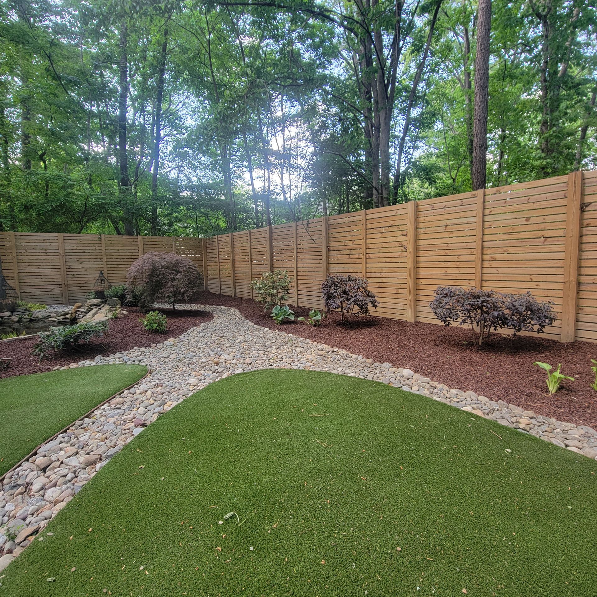 Landscaped backyard with green lawn, gravel path, mulch beds, shrubs, and a wooden privacy fence under trees