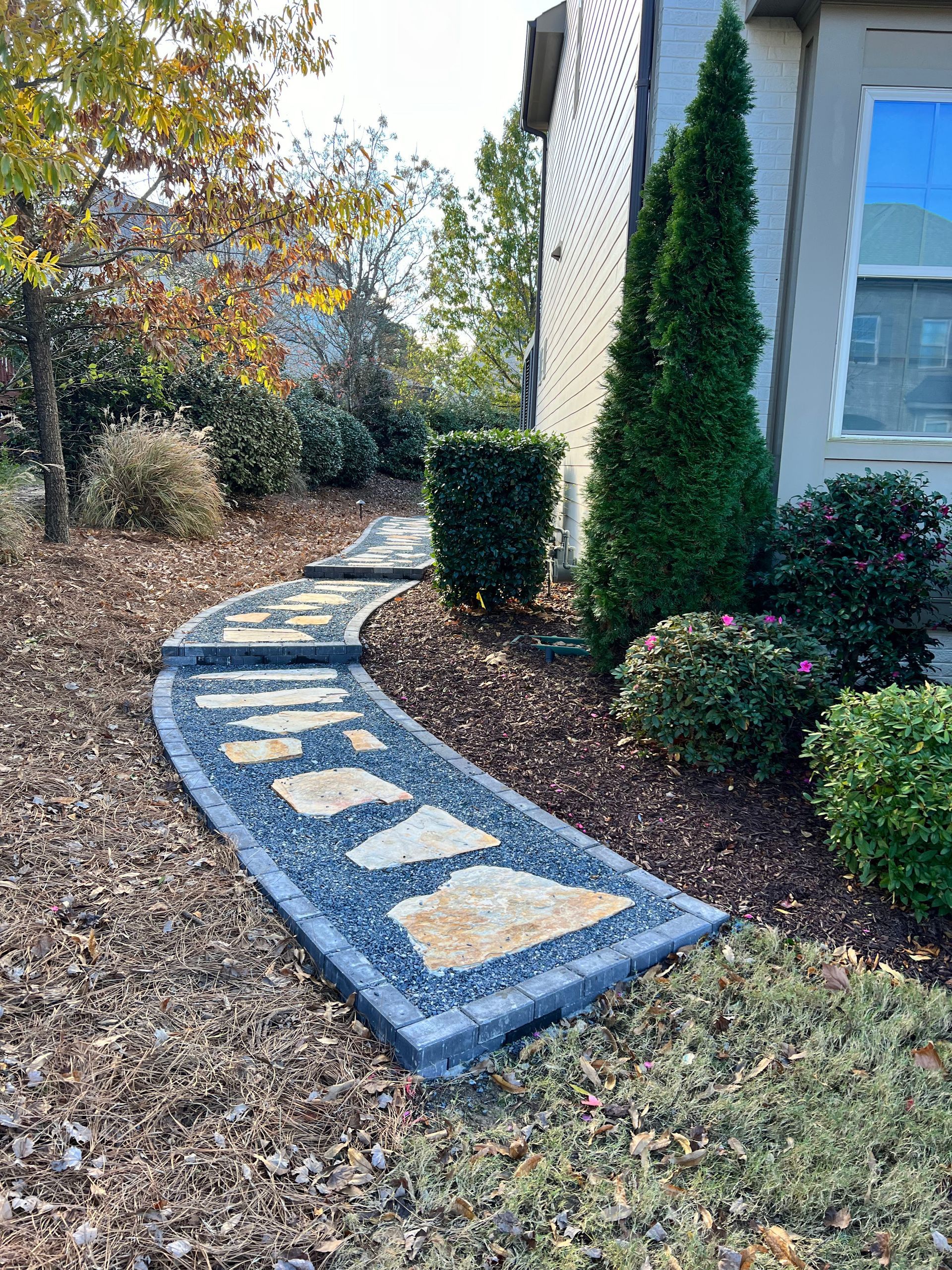 Curved stone garden path with mosaic stepping stones beside a house and shrubs in fall.