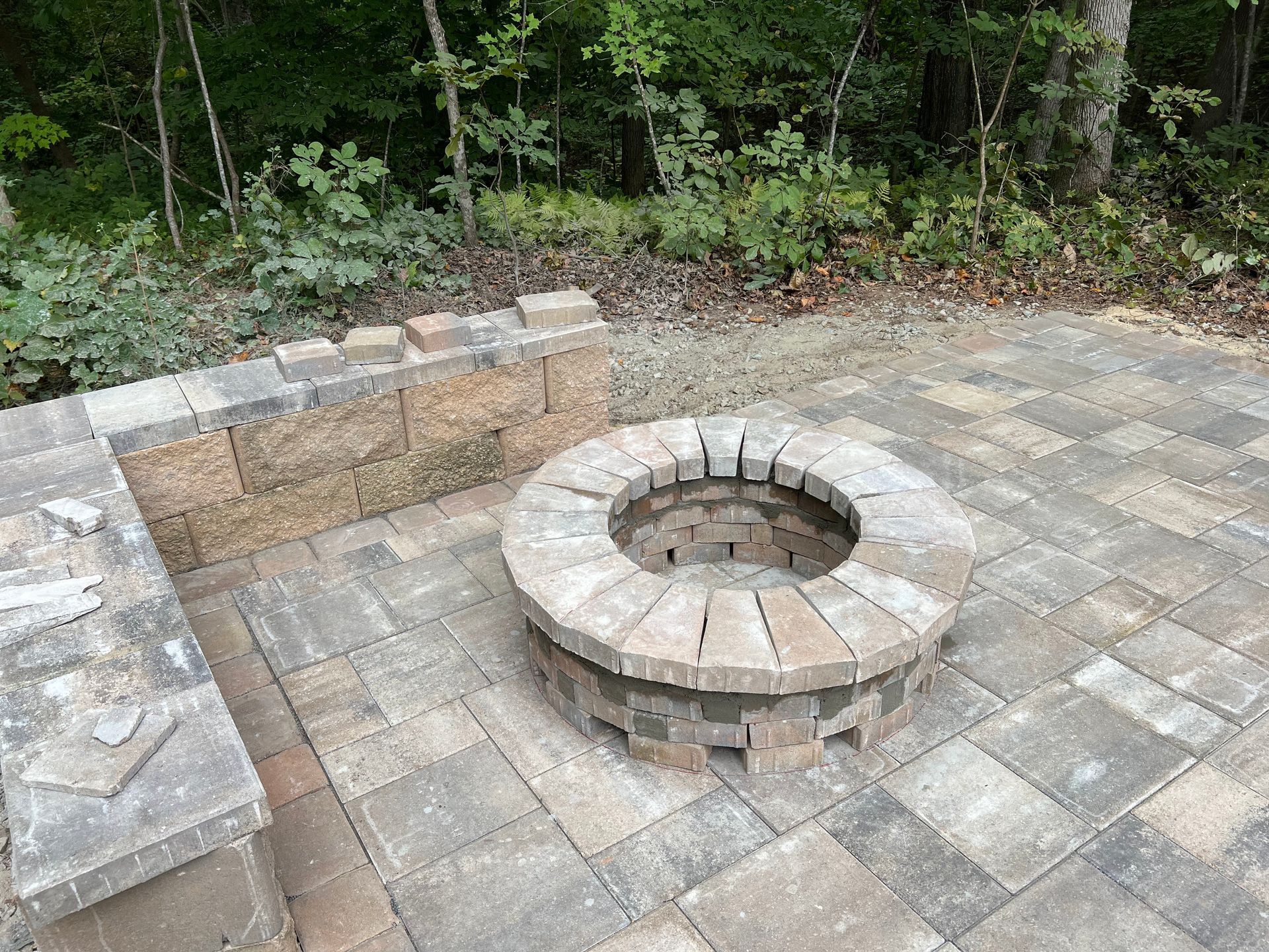 Stone fire pit and curved bench seating on a patio beside a wooded area.