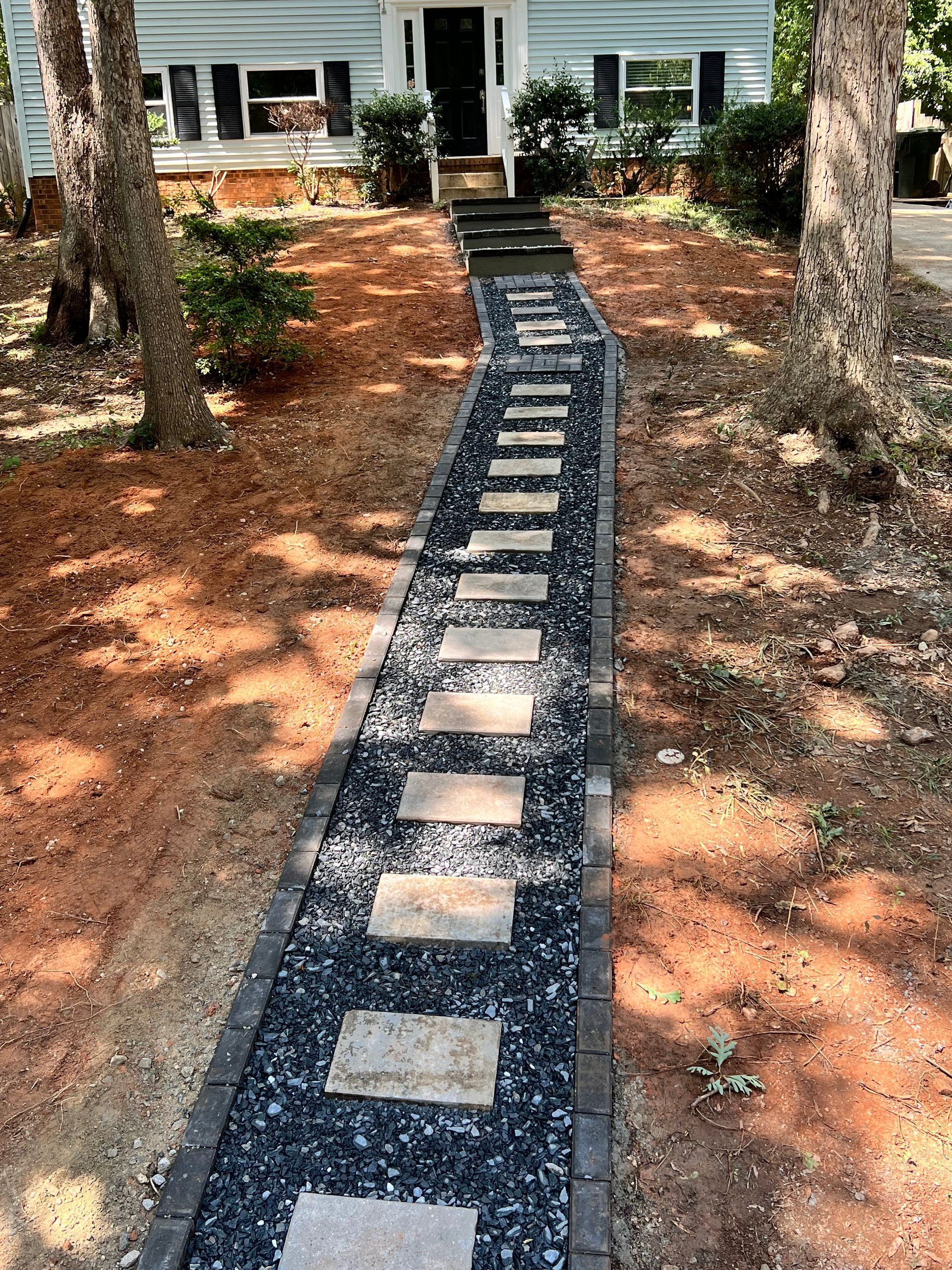 Stone stepping path leads through pine mulch to a house entrance between trees.