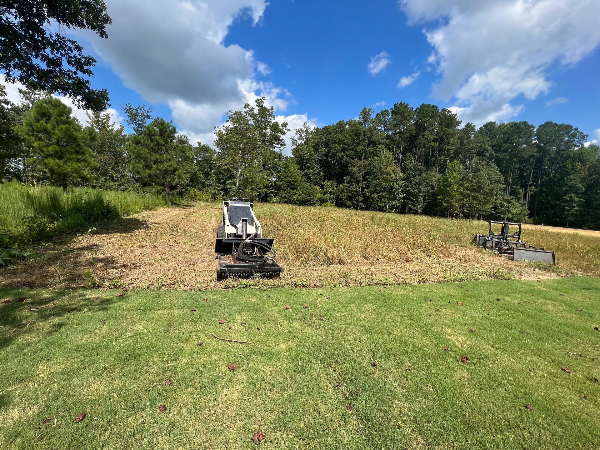 Two utility tractors on a grassy hillside under a blue sky, mowing or clearing a field.