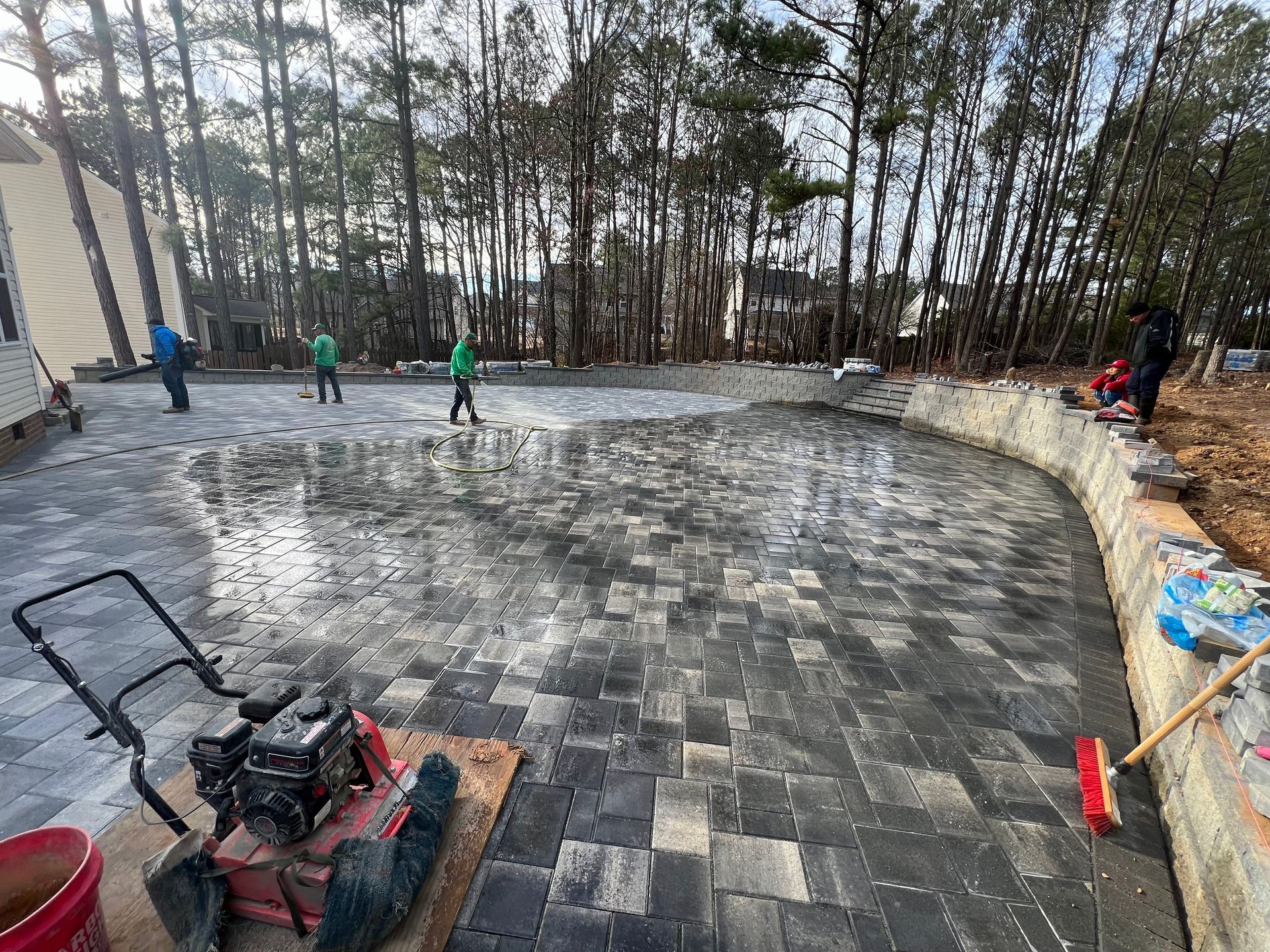 Workers paving a curved stone patio beside a house and wooded yard