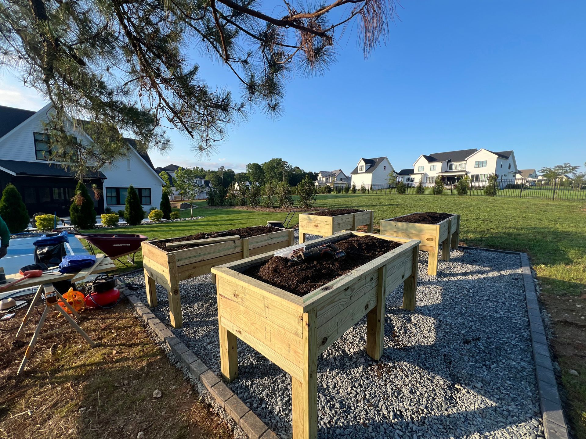 Raised wooden garden beds on a gravel yard with houses in the background under a blue sky