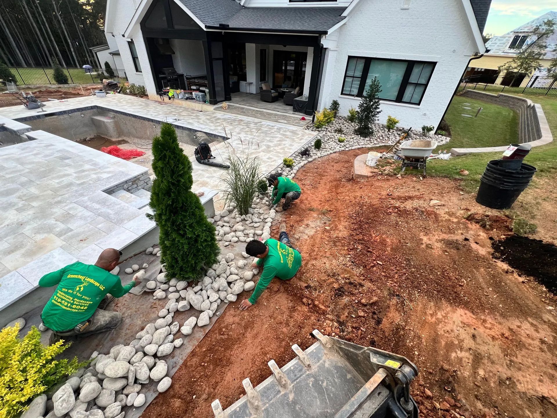 Workers installing landscaping in front of a white house, with stone edging, soil, and potted shrubs.