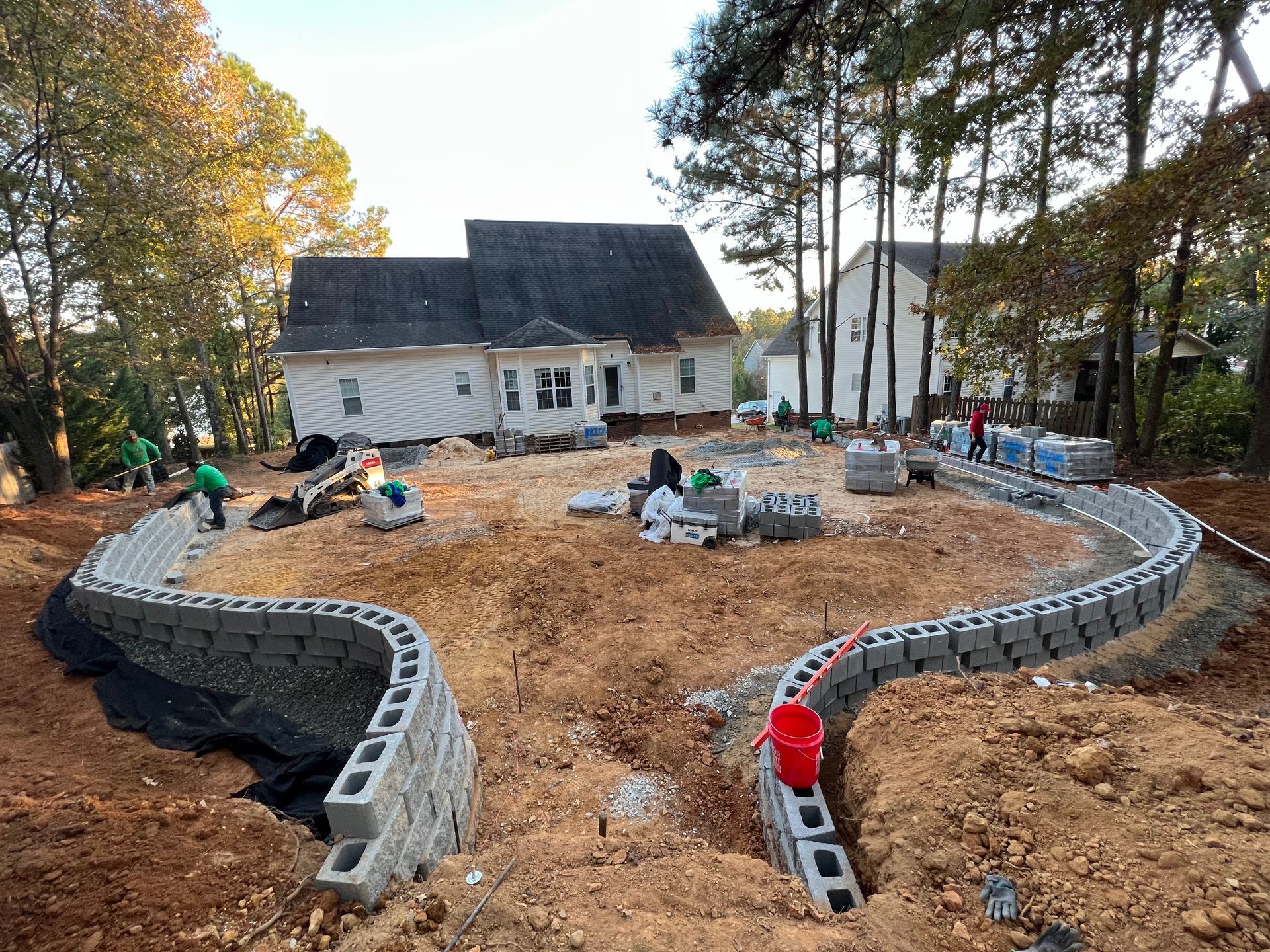 Workers building a curved cinder block border around a yard beside a house and trees