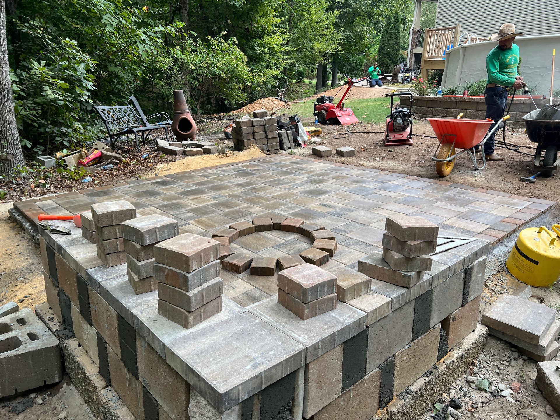 Outdoor brick patio construction with workers, stacked pavers, and a circular brick fire pit in progress