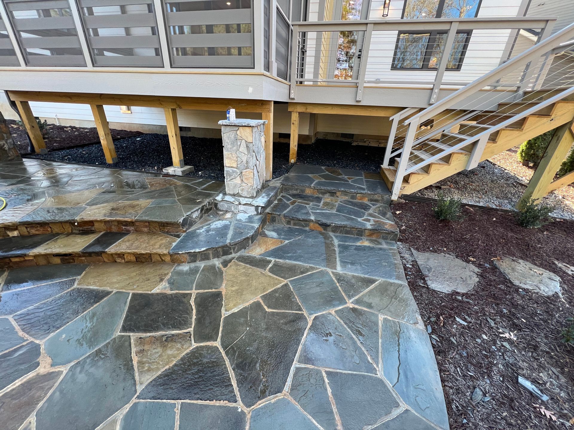 Stone patio under a raised wooden deck with stairs and a support post, showing a curved stone walkway.