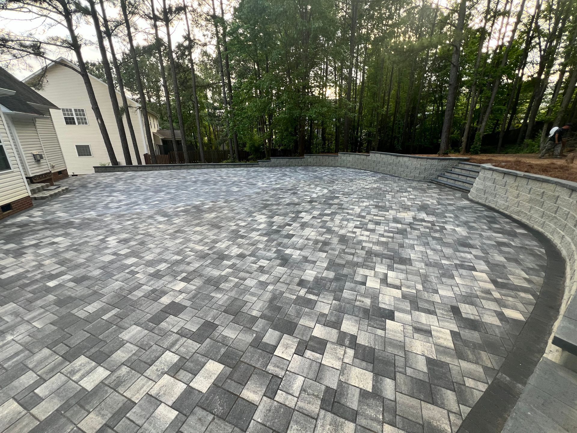 Curved gray paver driveway beside a house, bordered by trees and a low stone wall at dusk
