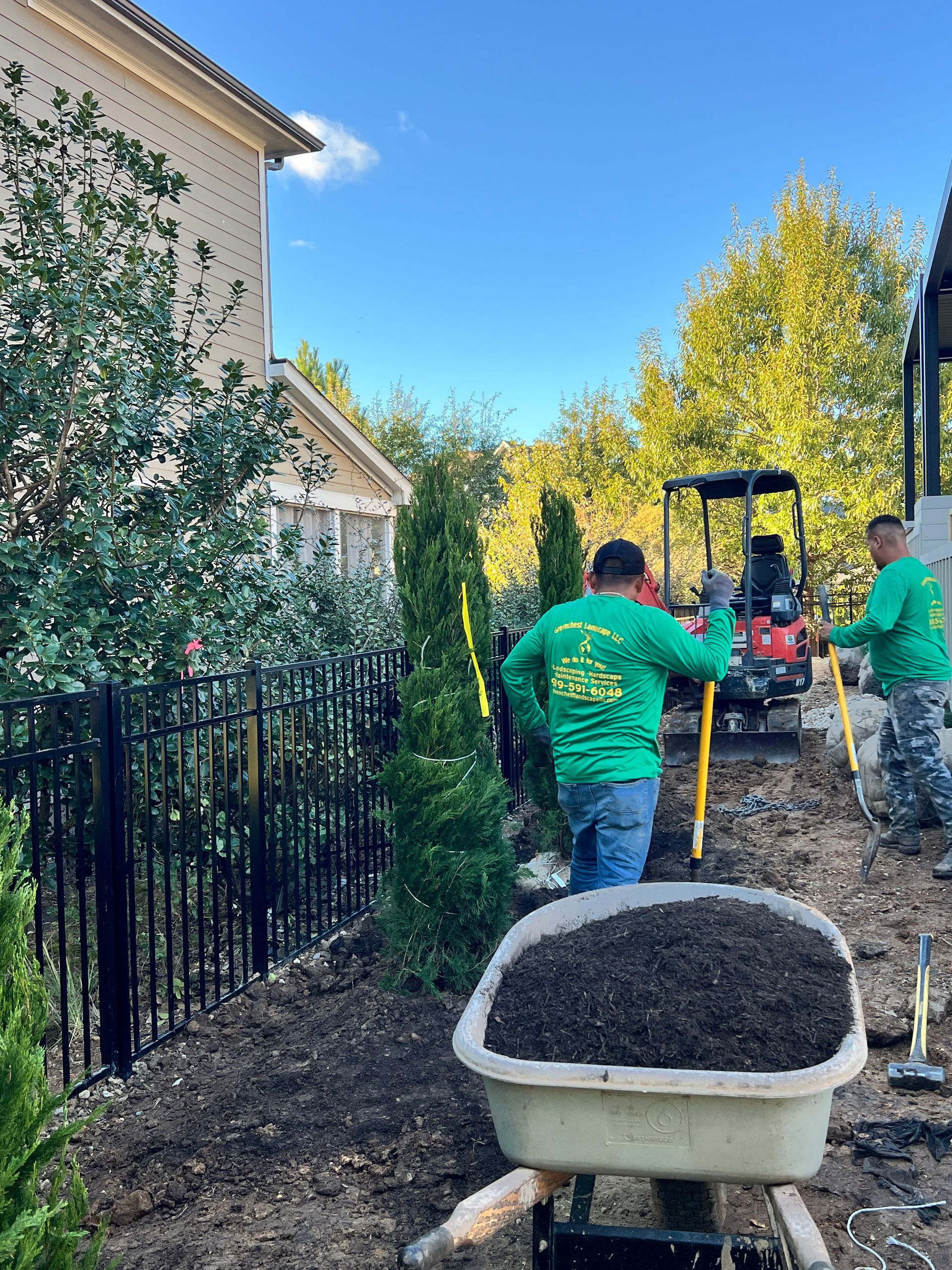 Workers moving soil with wheelbarrows along a fenced side yard beside a house and trees