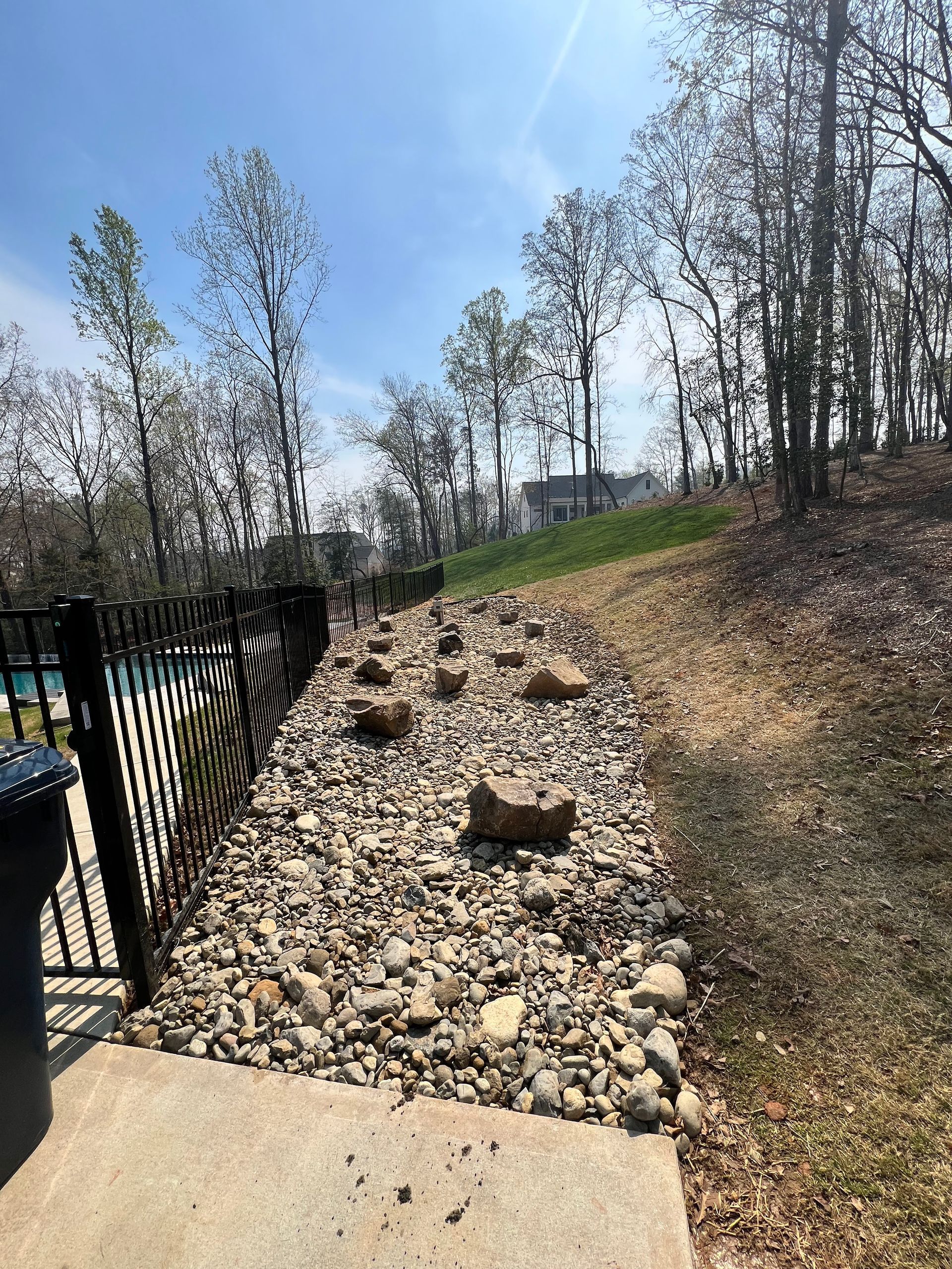 Rocky garden slope beside a black fence, with young trees and a concrete path in bright daylight