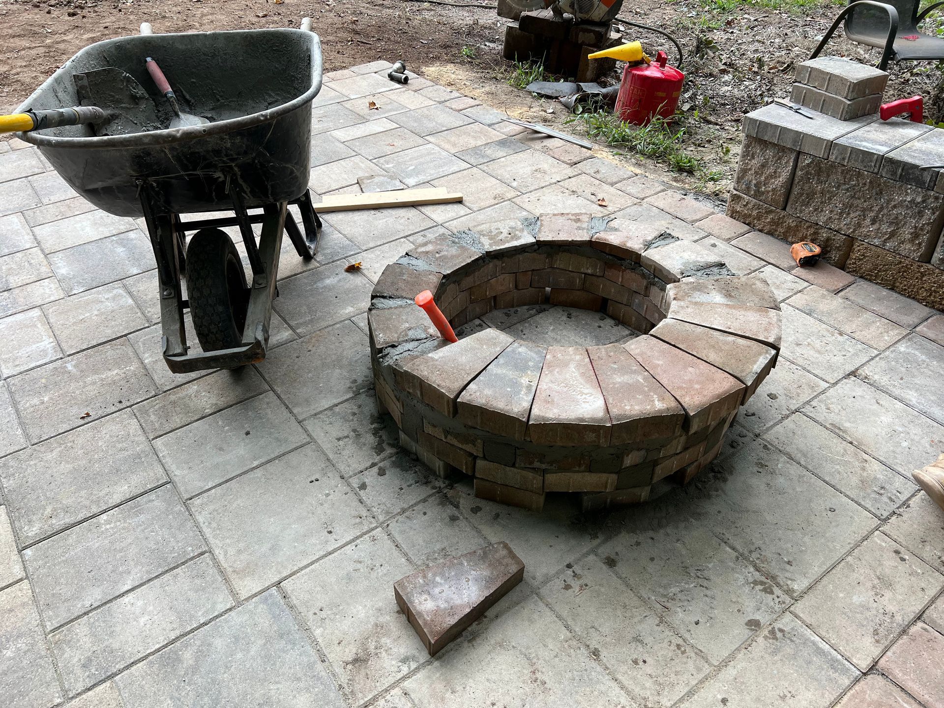Wheelbarrow beside a circular brick fire pit on a gray patio, with loose bricks and tools nearby