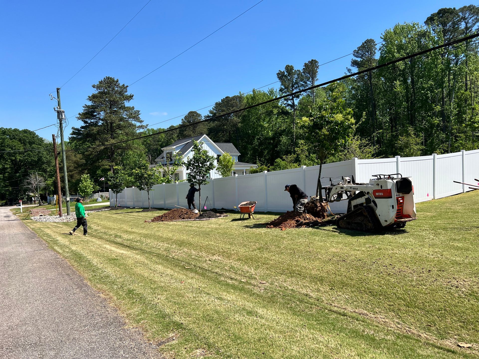 Workers trimming trees along a white fence beside a roadside, with a wood chipper in a grassy yard.