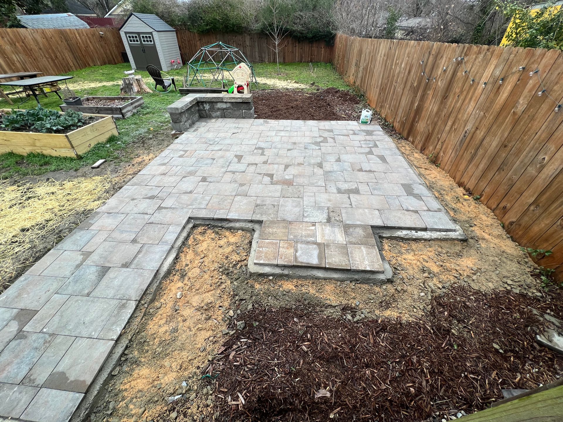 Backyard patio under construction with gray pavers, soil, and a wooden fence along the right side