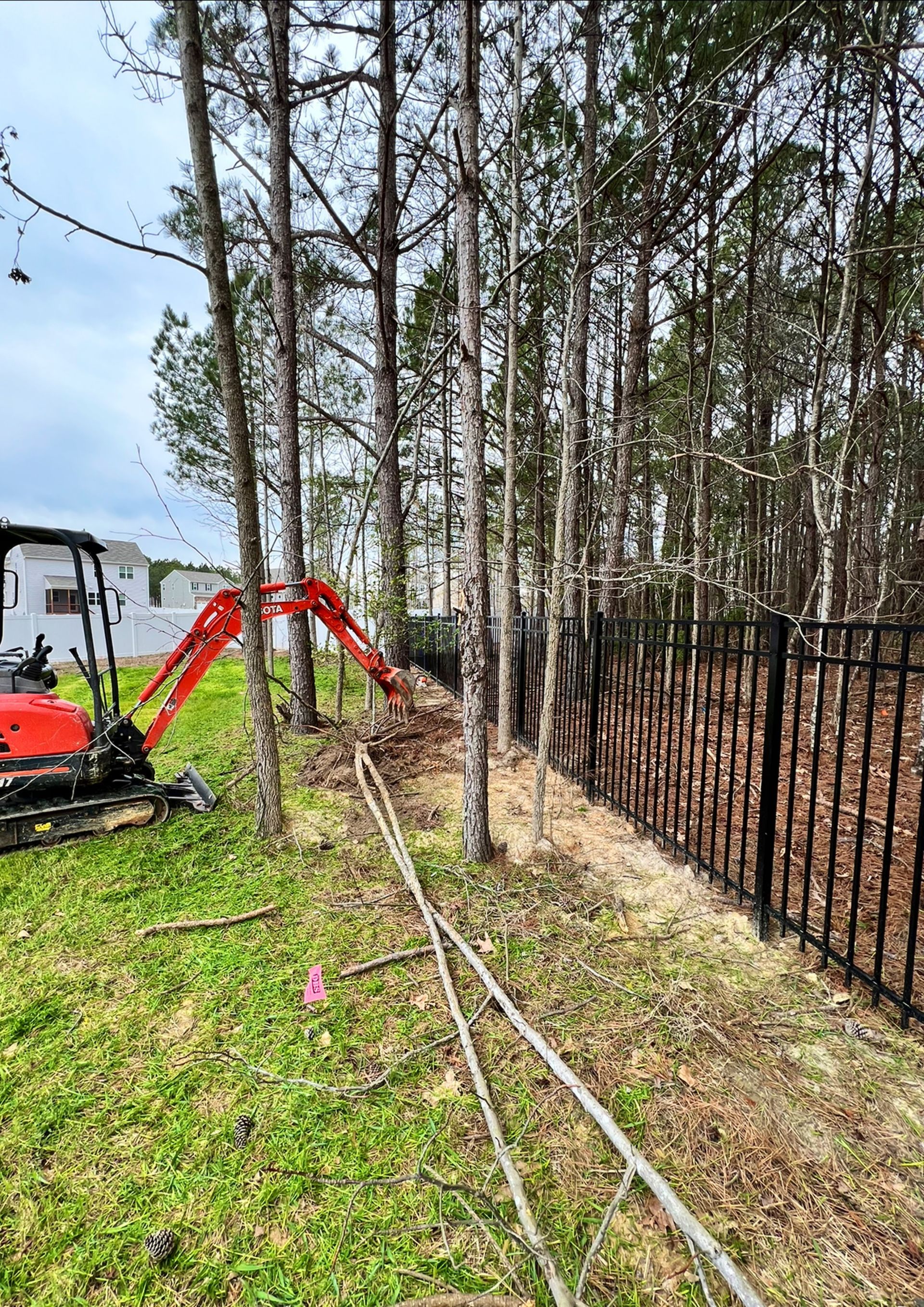 Small excavator beside a black fence, clearing brush in a wooded yard on green grass