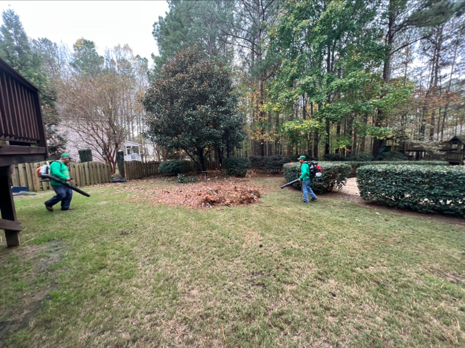 Two workers in green shirts clearing leaves in a wooded backyard near a house