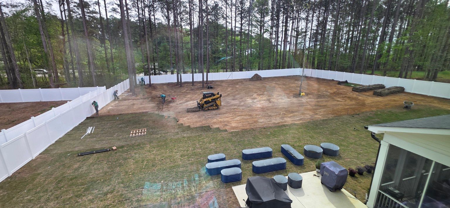 Wide backyard with a dirt construction area, excavator, white fence, stacked pavers, and trees beyond.