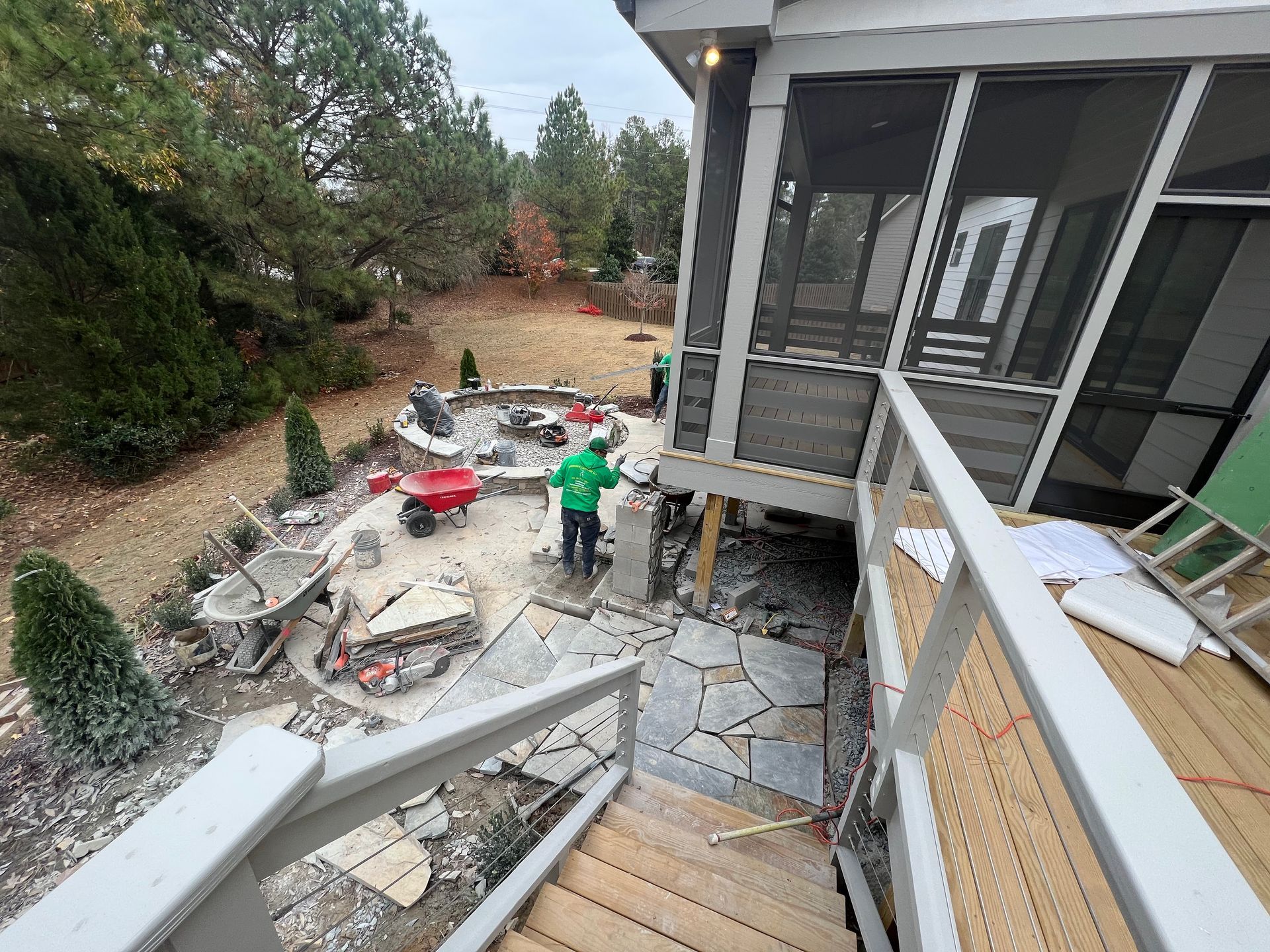 Backyard construction site with debris, stone patio, and a person in a green shirt near the deck