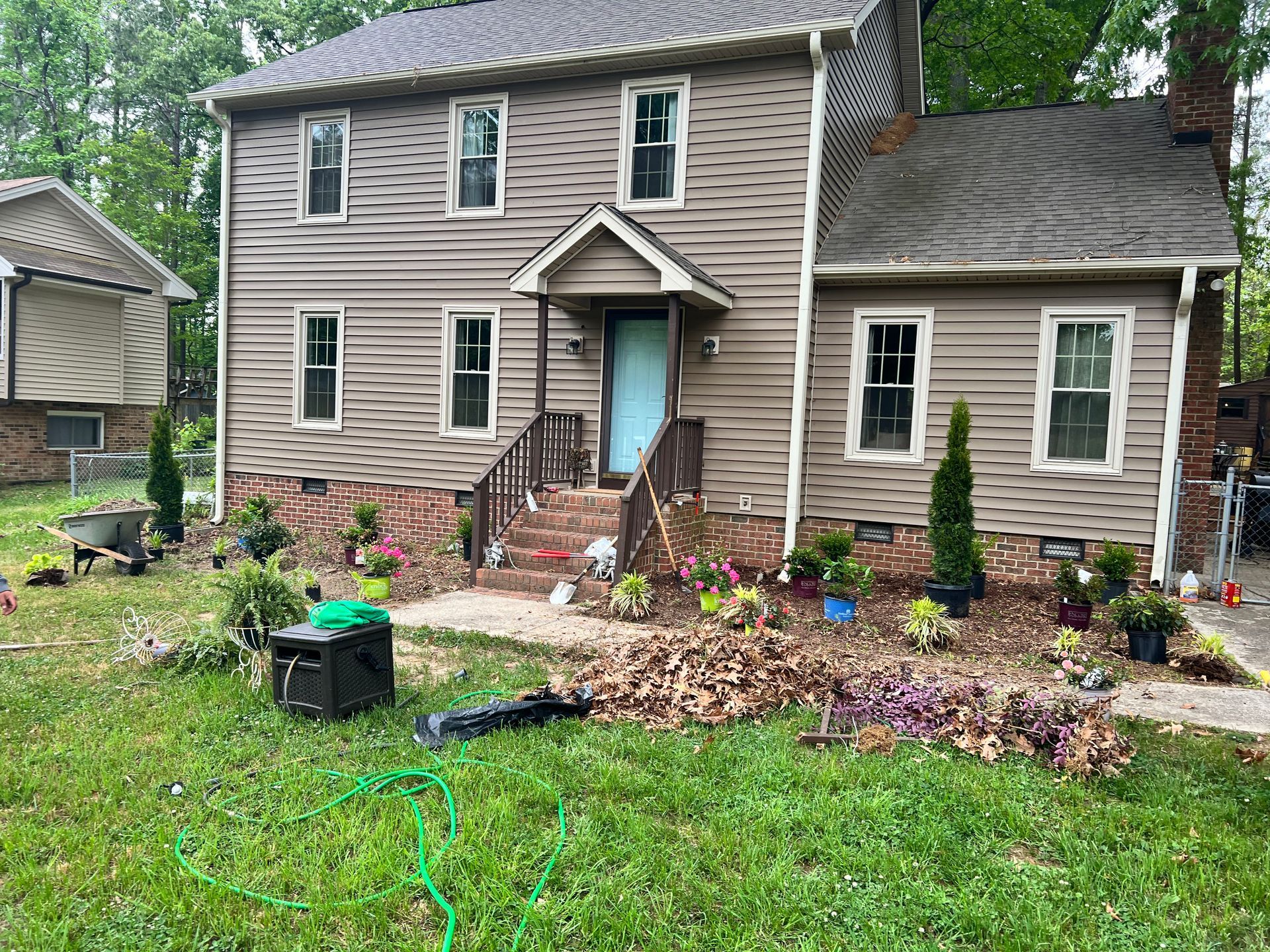Two-story beige house with stone steps, blue door, and a landscaped front yard with potted plants and garden hose