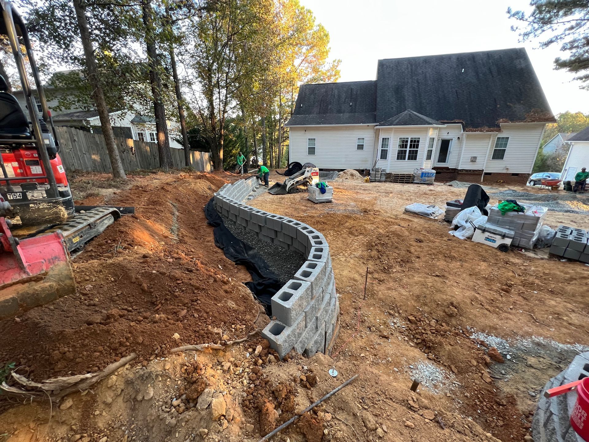 Construction site with curved cinder block wall, workers, and a white house in the background