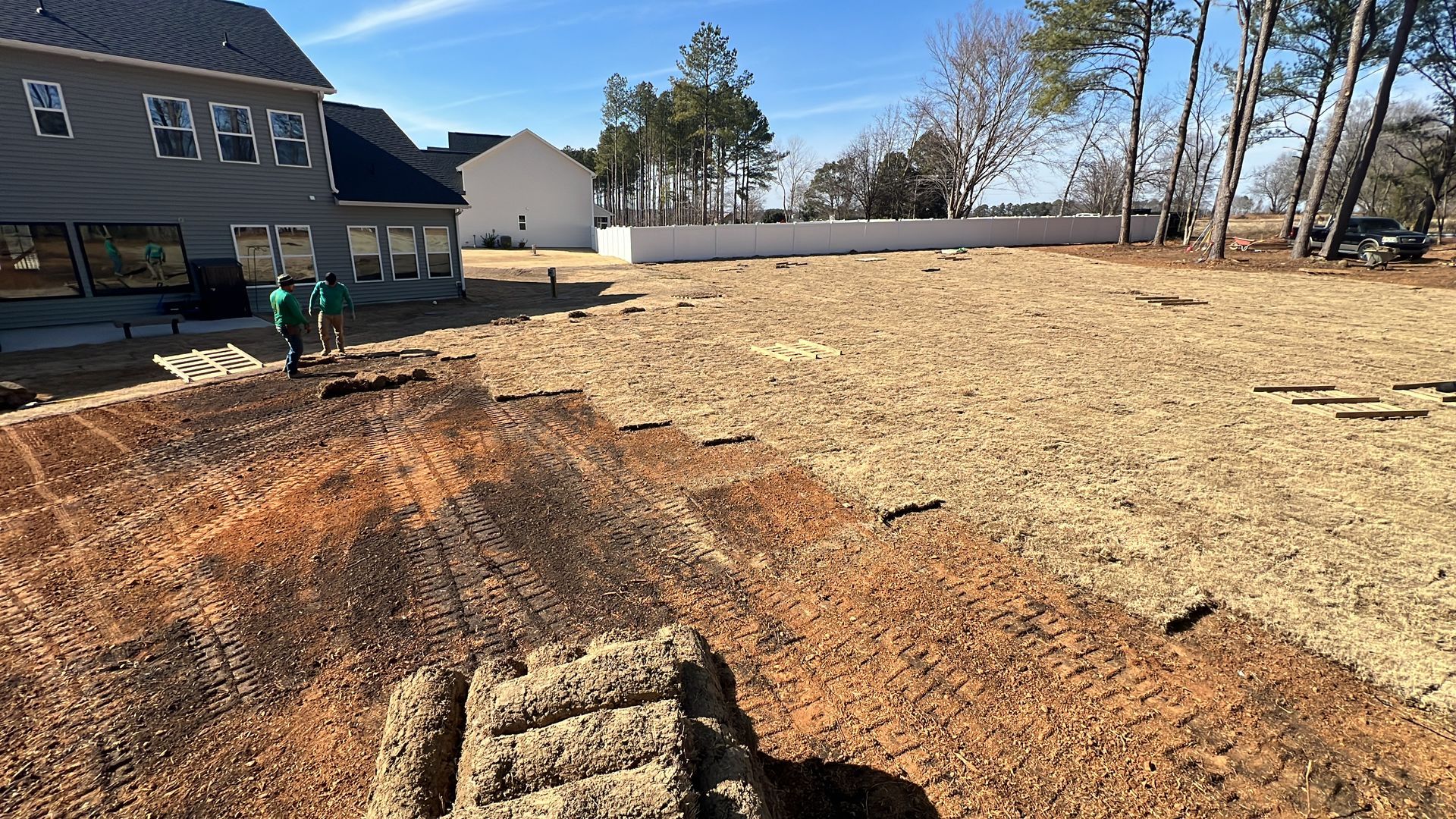 Large cleared dirt lot beside a house, with excavator tracks and a trench in the foreground under a clear sky