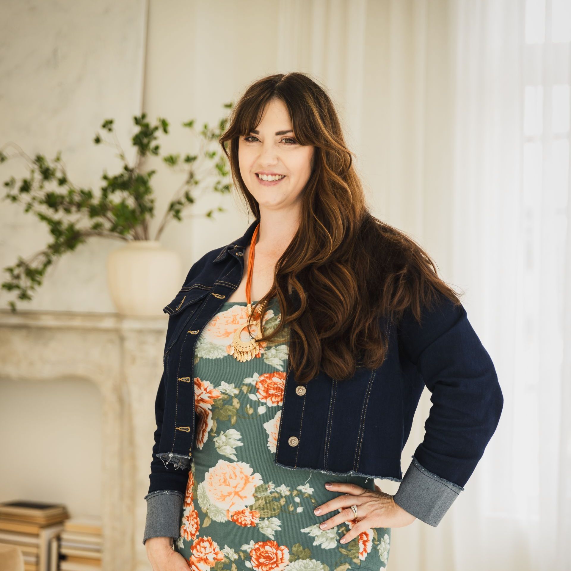 A woman wearing a floral dress and a denim jacket smiles for the camera