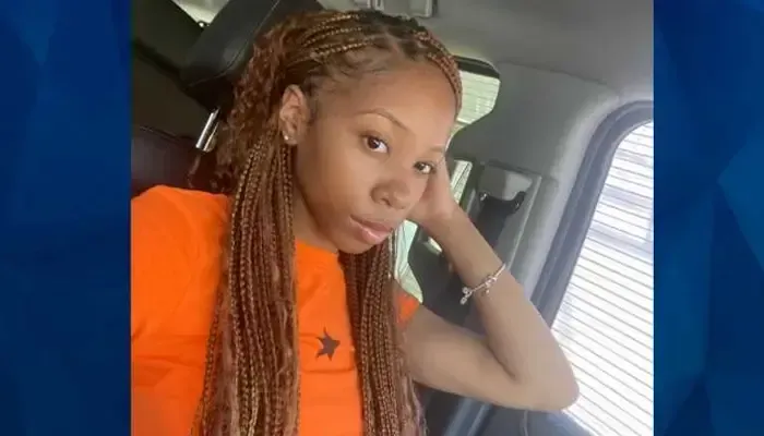 A woman with braids is sitting in a car with her hand on her head.
