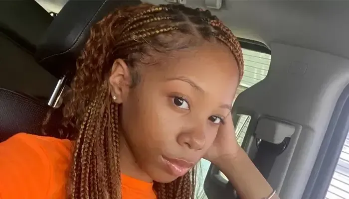 A woman with braids is sitting in a car with her hand on her face.