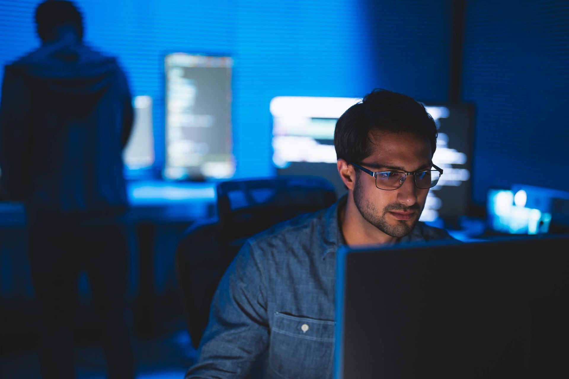 Two people working on computers in a dimly lit room with blue lighting and code on screens.