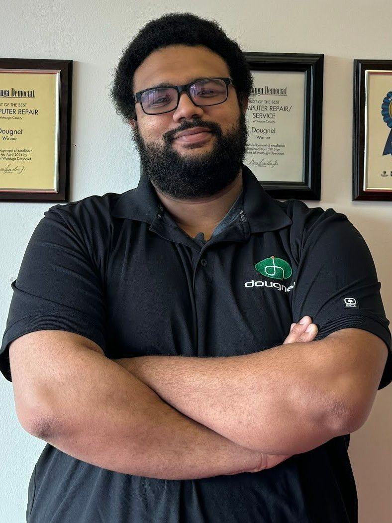 Man with beard and glasses, arms crossed, wearing black shirt, smiling in front of framed documents.