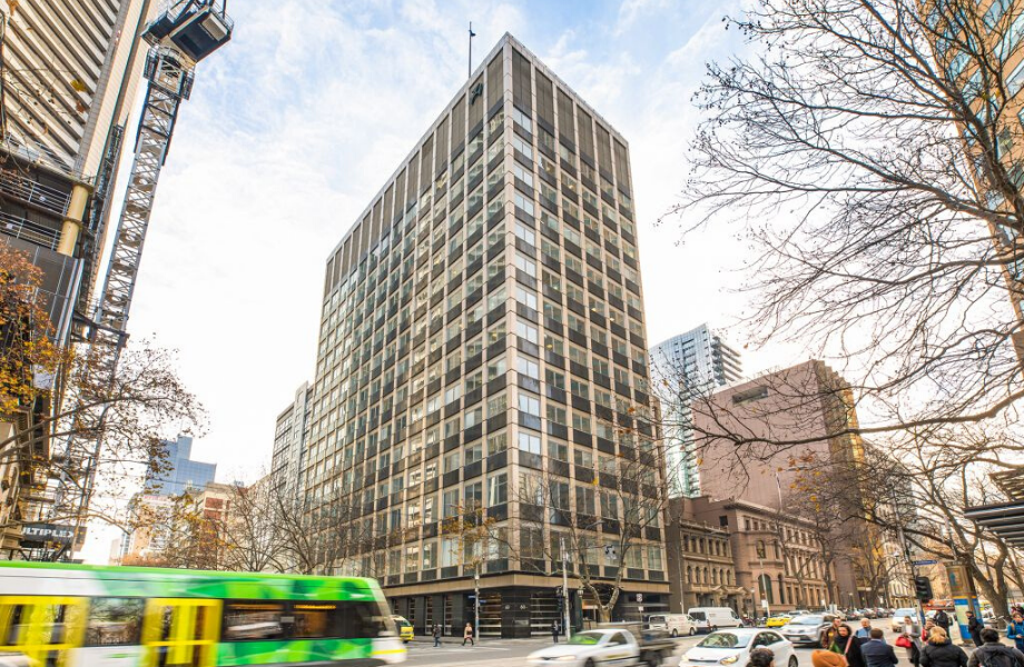 A green and white bus is driving down a city street in front of a tall building.