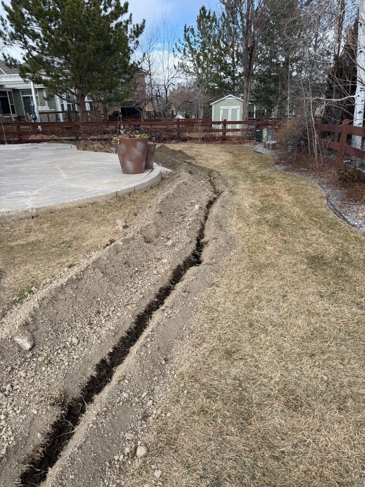 A narrow trench dug through a grassy yard alongside a stone patio and a wooden fence.