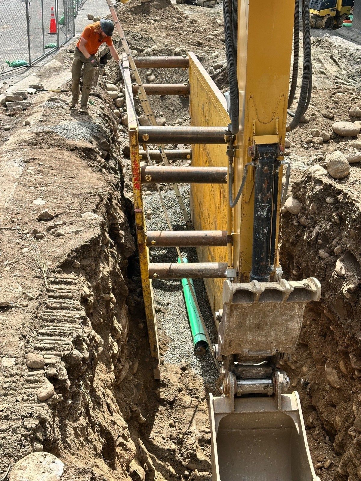 A worker in a high-visibility orange shirt stands by a trench-box shield in an excavated construction site.