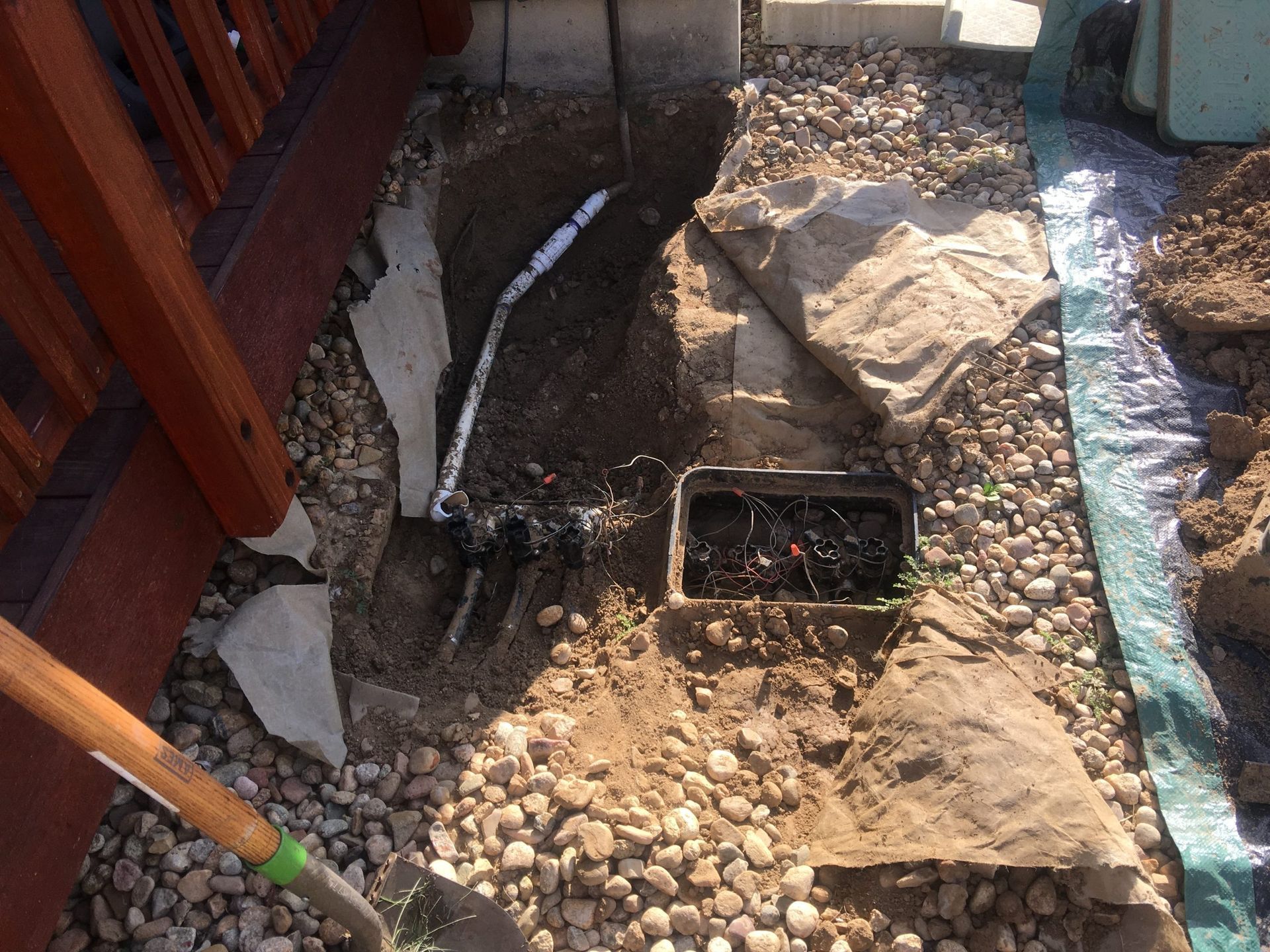 An open trench in a rocky yard shows exposed white PVC piping connected to an irrigation box near a wooden deck.